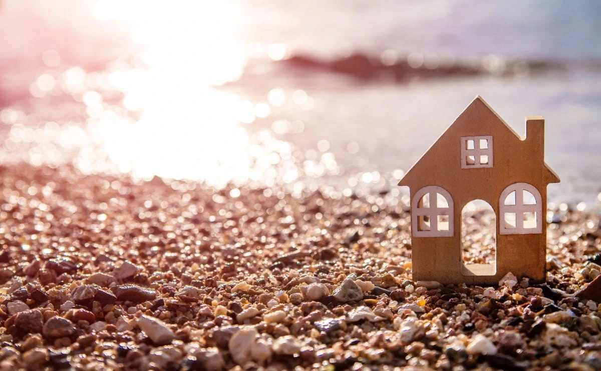 Casa de madera en miniatura sobre la arena de una playa con el sol reflejado en el agua