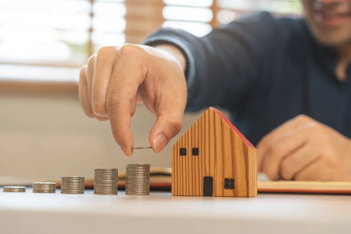 Mano colocando una moneda sobre pilas de monedas junto a una casa de madera en miniatura, como símbolo de ahorro e inversión inmobiliaria