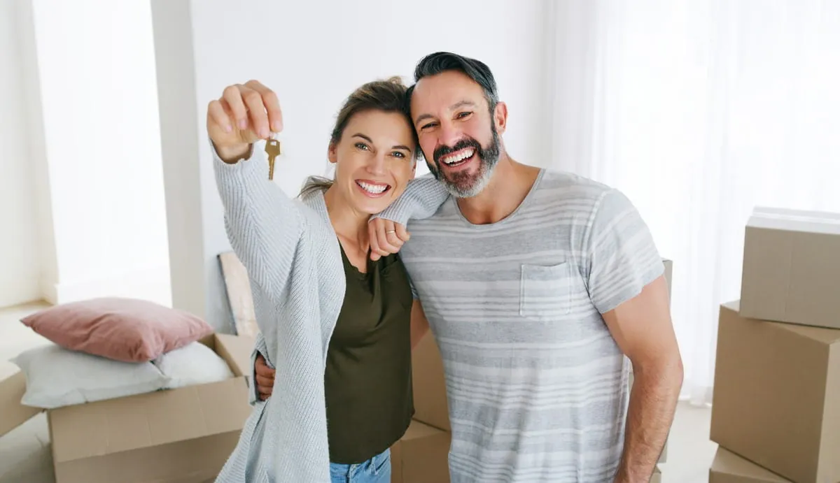 Pareja sonriente mostrando las llaves de su nueva propiedad entre cajas de mudanza