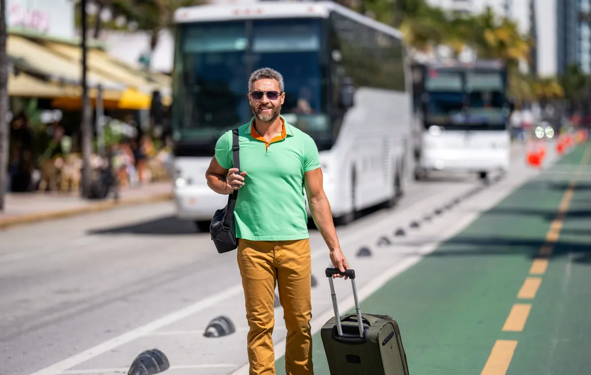 Hombre con gafas de sol y maleta de viaje caminando por una avenida de Miami Beach, con buses al fondo