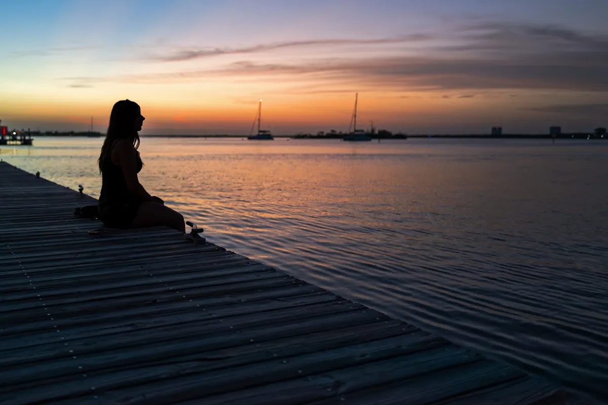 Silueta de una mujer sentada en un muelle de madera frente al agua al atardecer, con veleros al fondo
