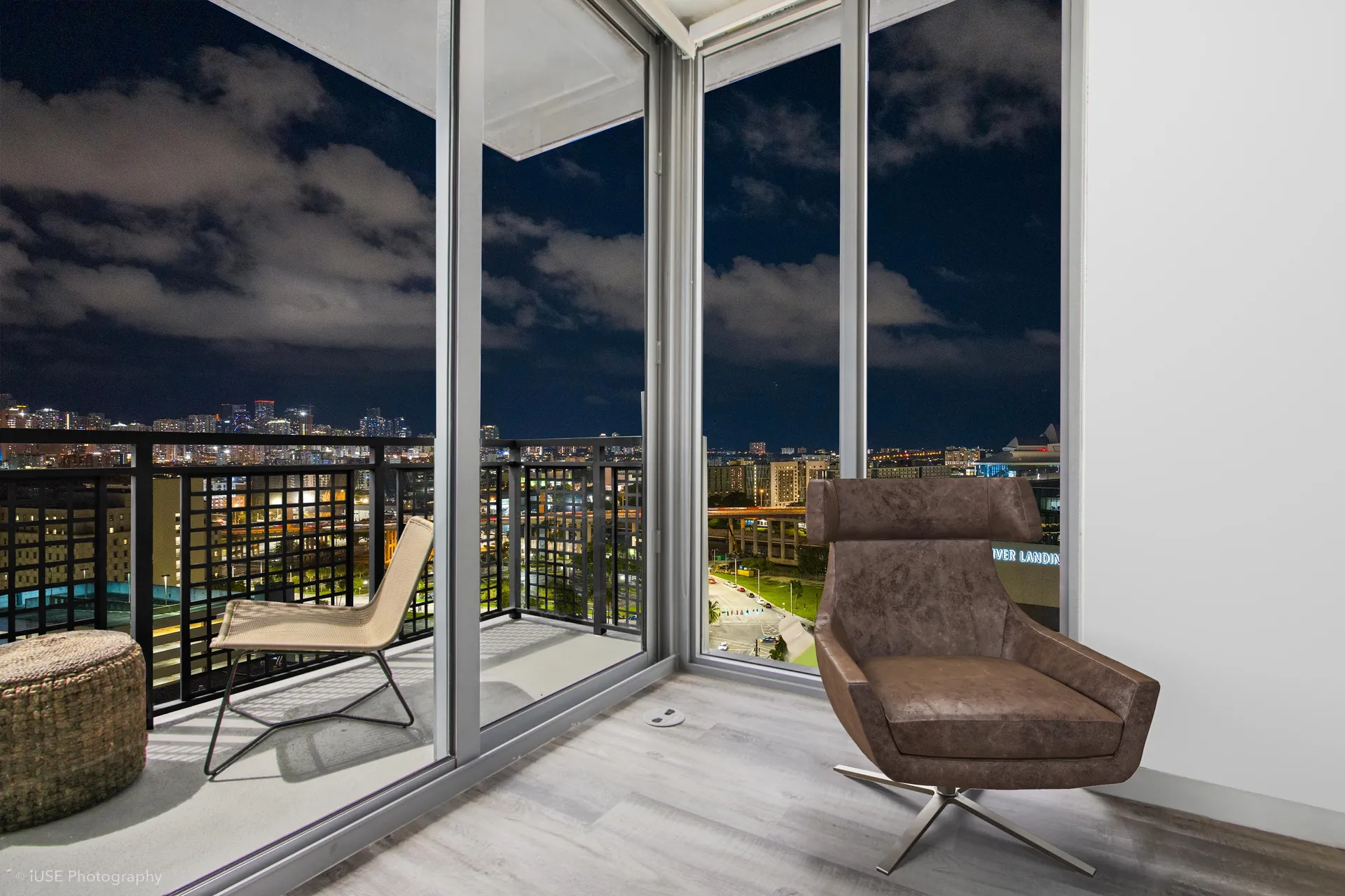 River District 14 — sala de una residencia al anochecer, con ventanal de piso a techo, butaca de cuero y salida al balcón con las luces de la ciudad al fondo