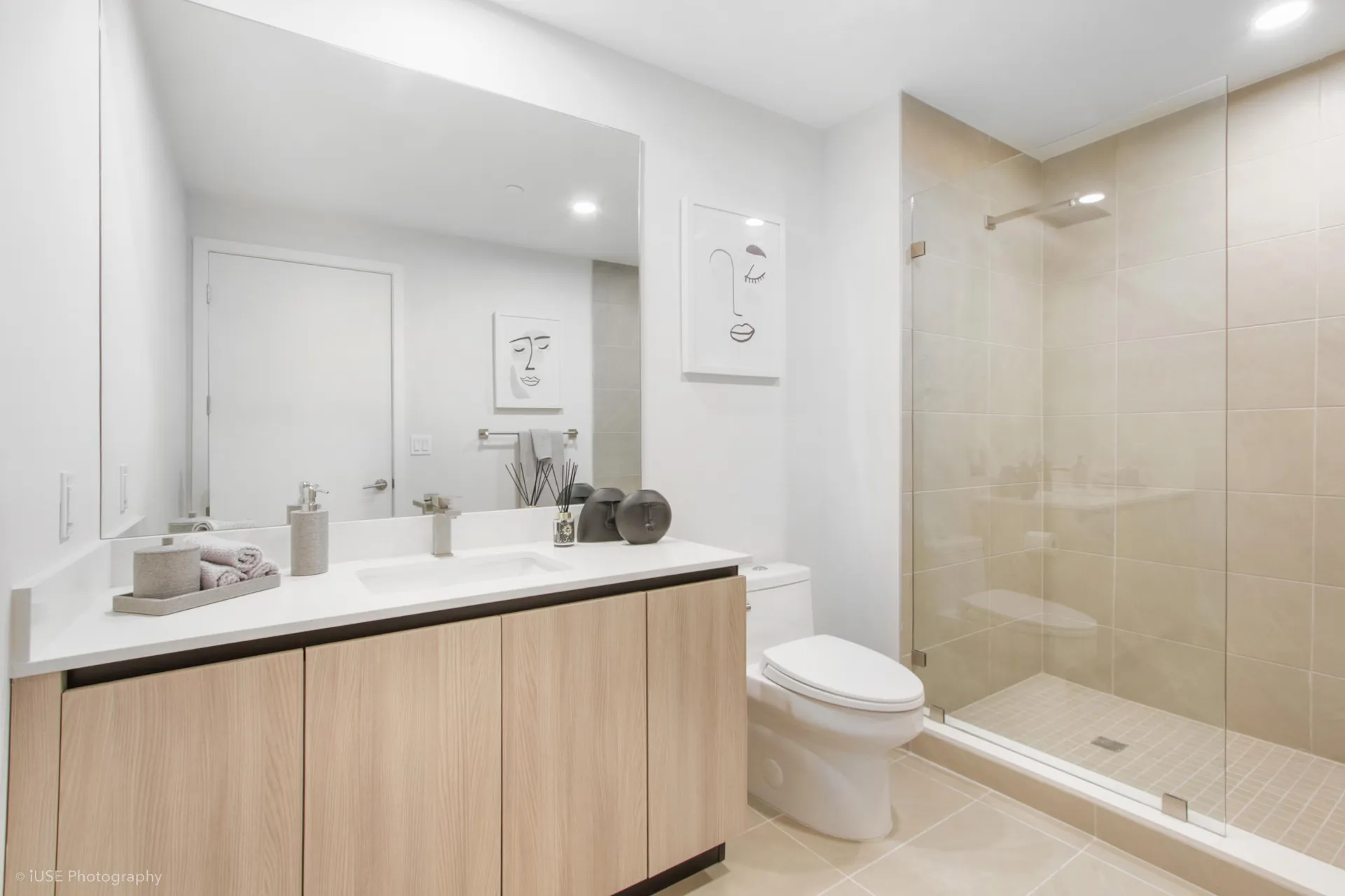 River District 14 — baño de una residencia con mueble de madera clara, cubierta blanca, espejo iluminado y ducha de lluvia con mampara de vidrio