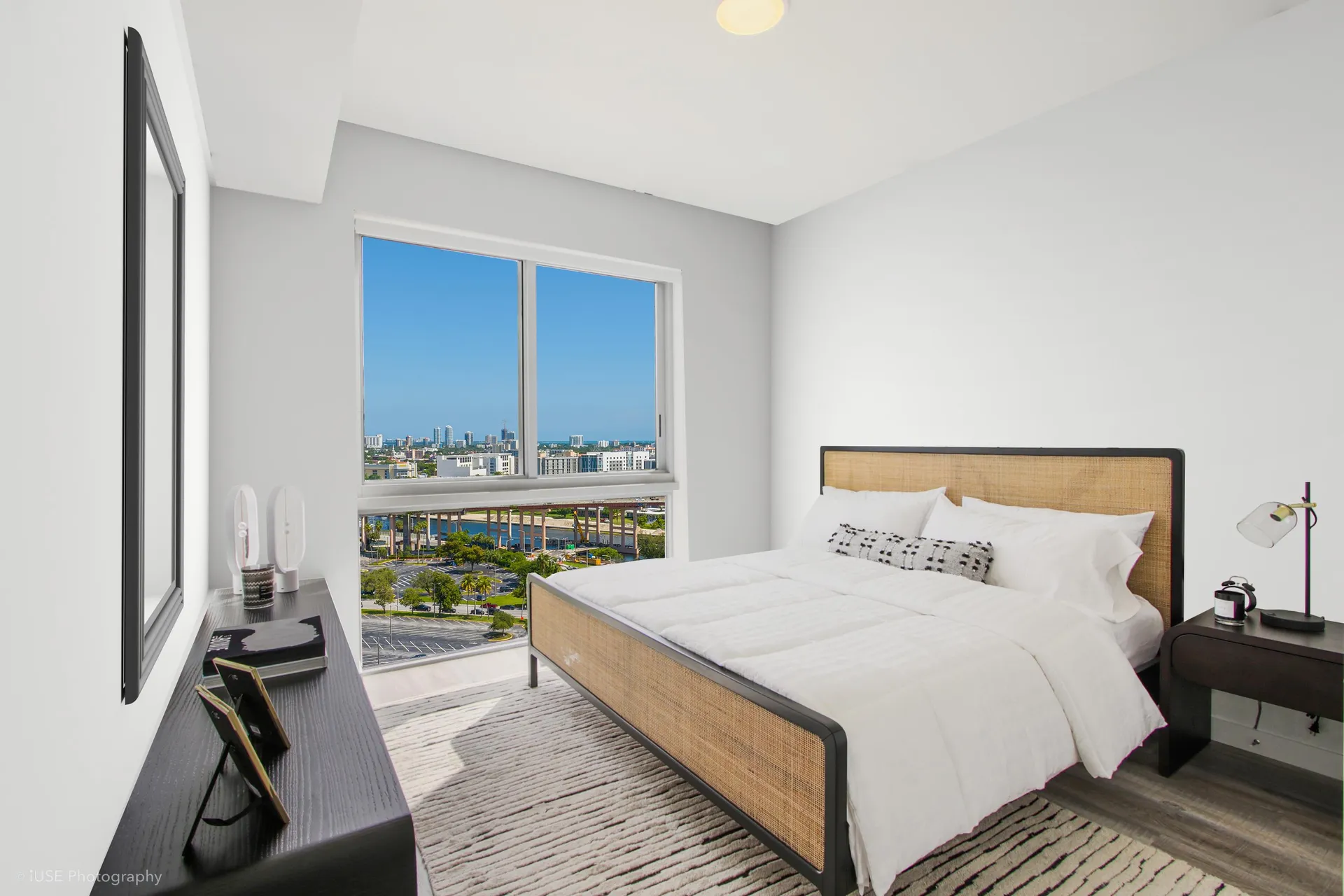 River District 14 — recámara principal amoblada con cabecera de madera, ropa de cama blanca y ventana con vista abierta hacia la ciudad
